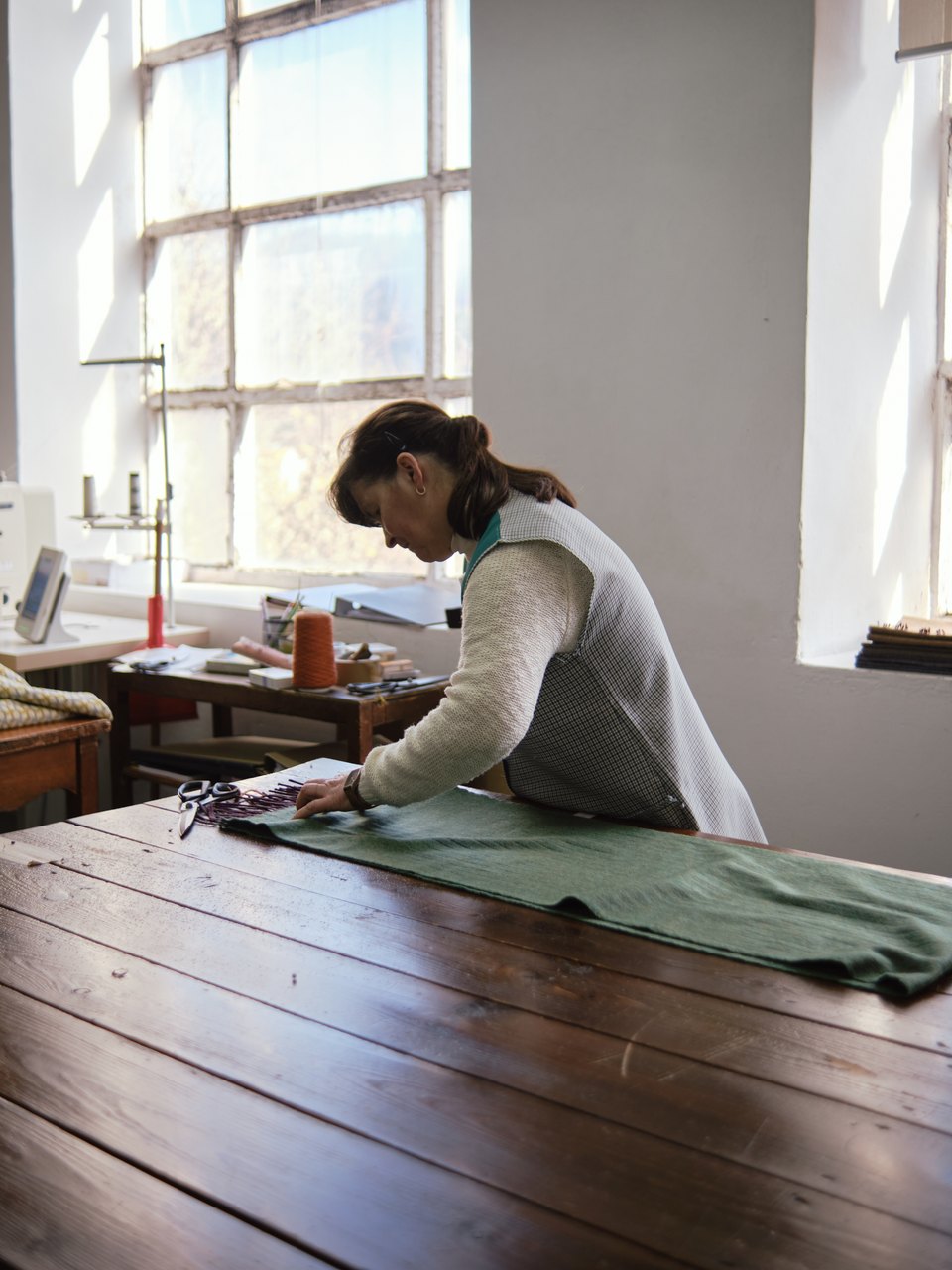 Burel is a versatile fabric used in coats, wall panels, and accessories, crafted in a workshop filled with natural light. In this photo, a woman carefully aligns the fringes of a wool scarf. While this fabric has not undergone the treatment that defines burel, it is made from the same wool. Photo taken at Burel Factory